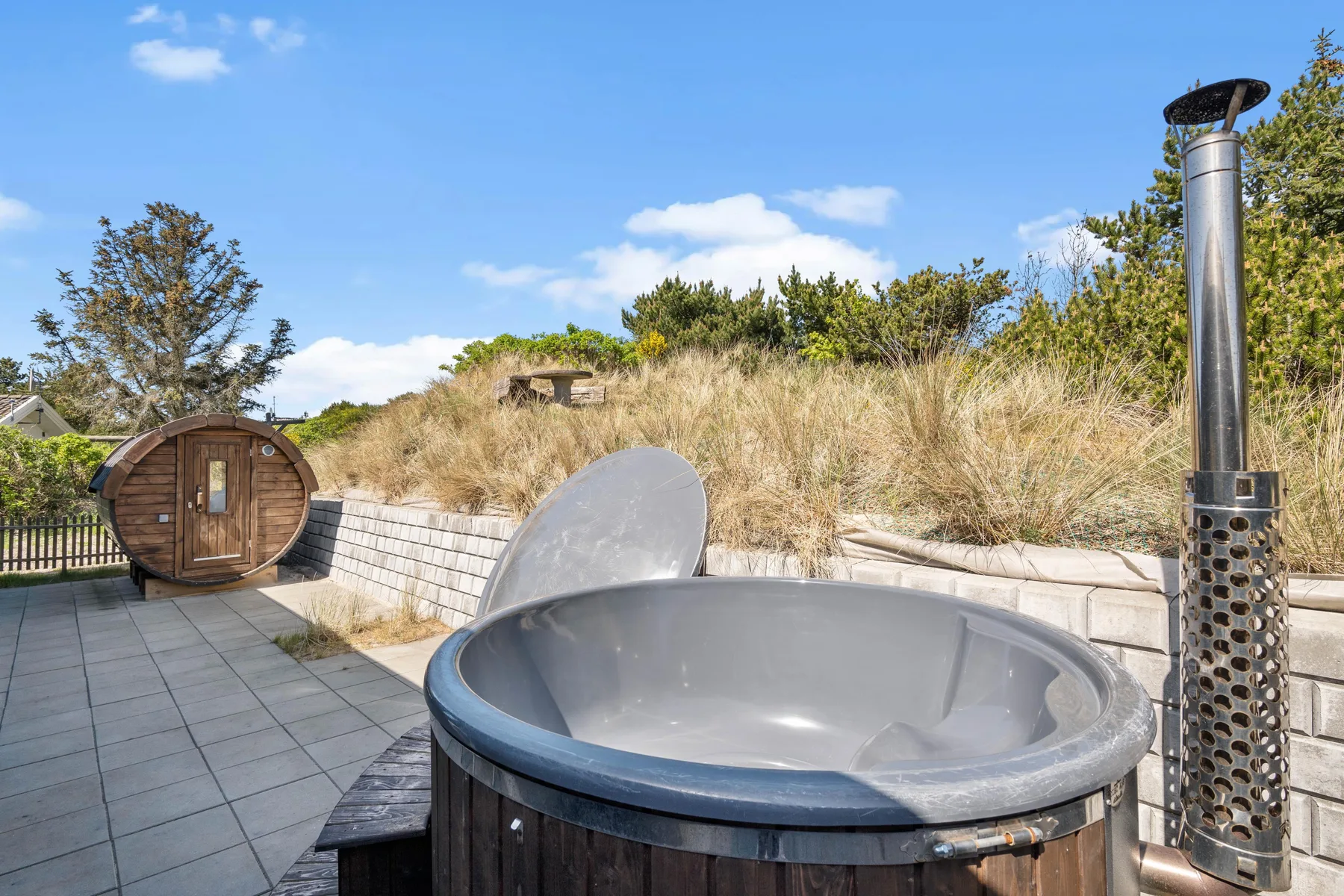 Una bañera de hidromasaje exterior está en un patio pavimentado junto a una sauna de madera.