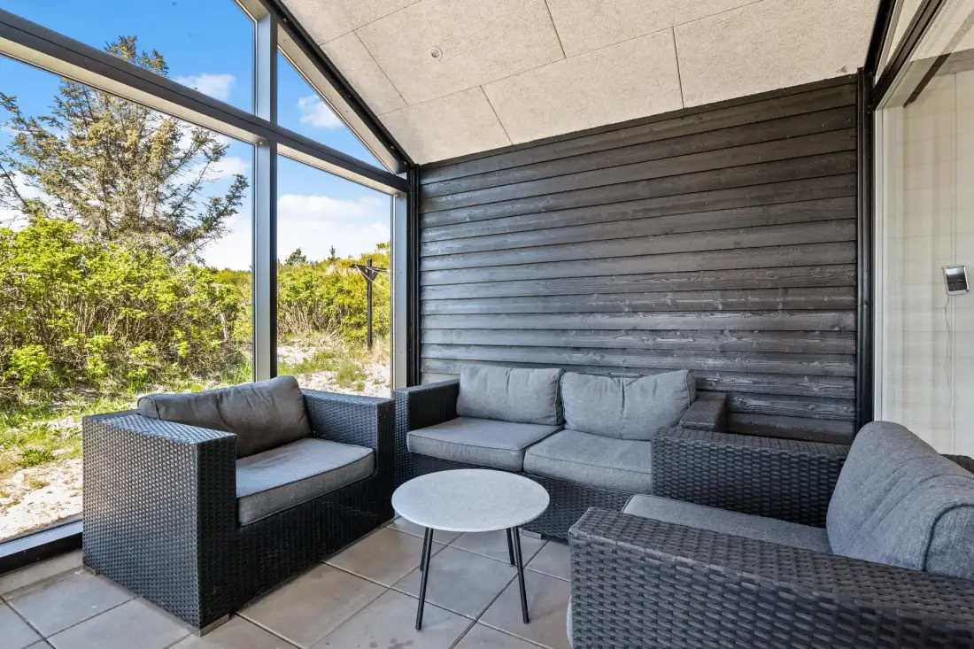 Una luminosa zona de estar cerrada tiene grandes ventanas y muebles tejidos oscuros.