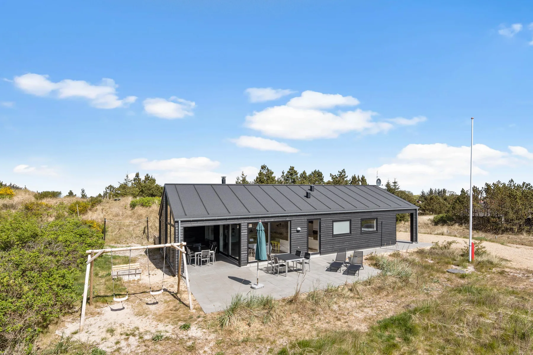 Una casa de vacaciones de fachada oscura se alza entre dunas arenosas y vegetación baja.