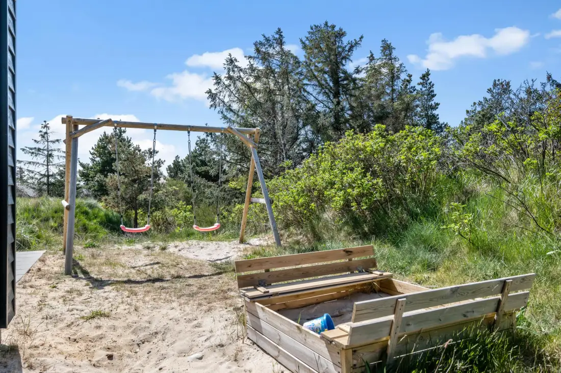 Una zona de juegos exterior de arena tiene columpios y un arenero de madera, rodeados de vegetación.