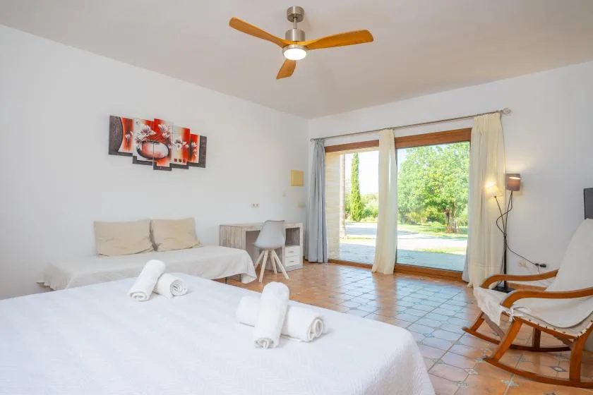 Un dormitorio luminoso con camas, ventilador de techo y puertas de vidrio hacia el jardín.