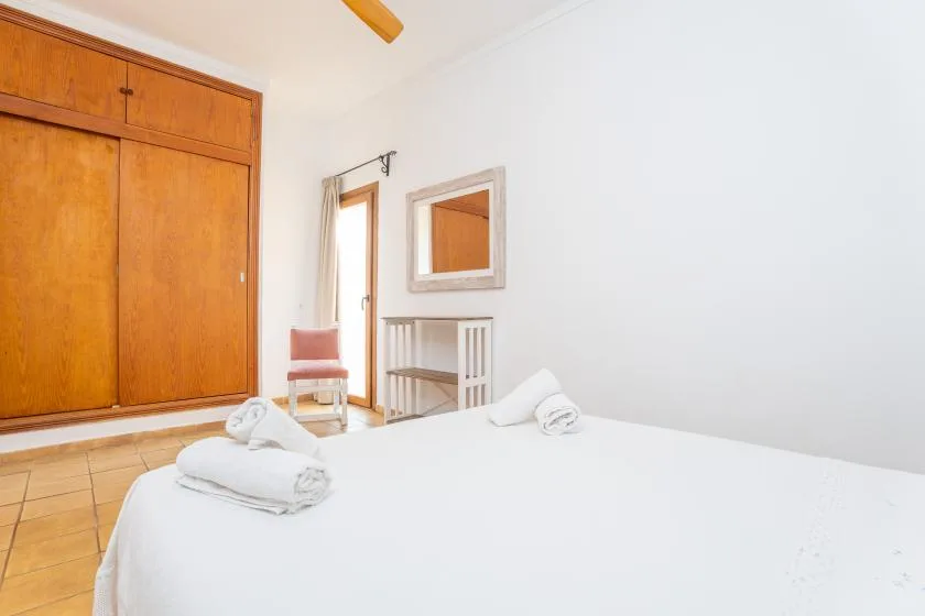 Un dormitorio luminoso con cama blanca, armario de madera y una pequeña zona de estar.