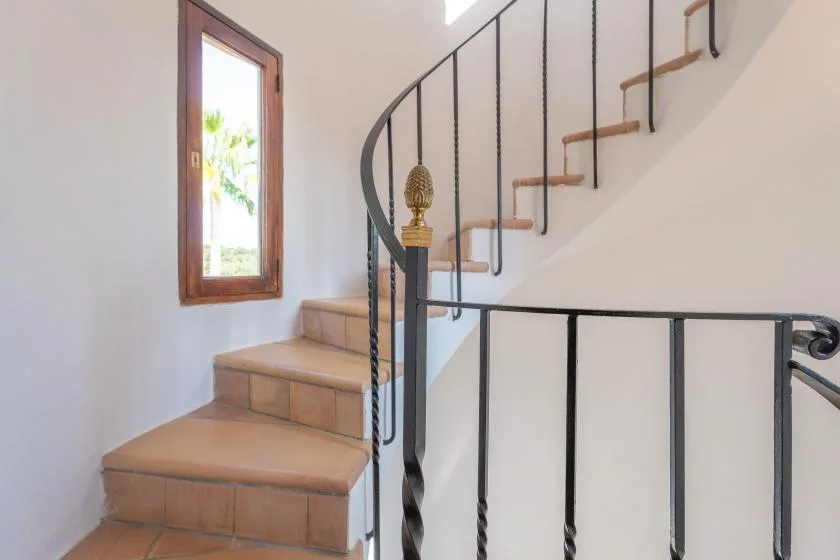 Una escalera de azulejos sube junto a una ventana estrecha y una barandilla negra.