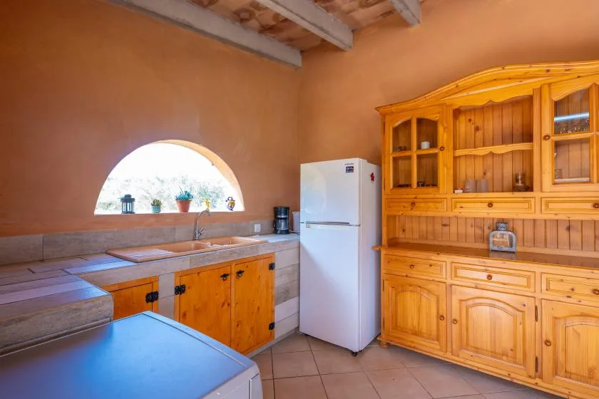 Una cocina rústica con gabinetes de madera, un refrigerador y un fregadero bajo una ventana arqueada.