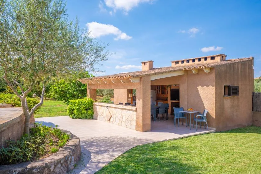 Una zona de comedor exterior a la sombra junto a un edificio de estuco, con vistas al jardín.