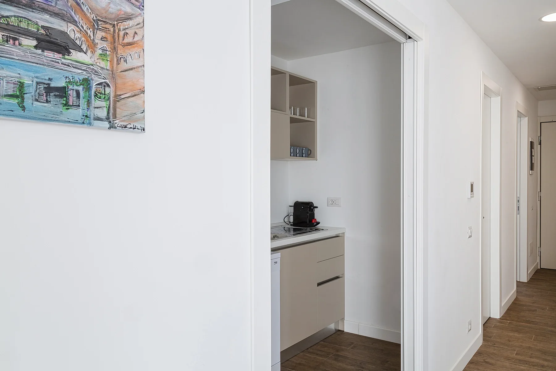A bright hallway opens onto a compact kitchenette with cabinets and a coffee maker.