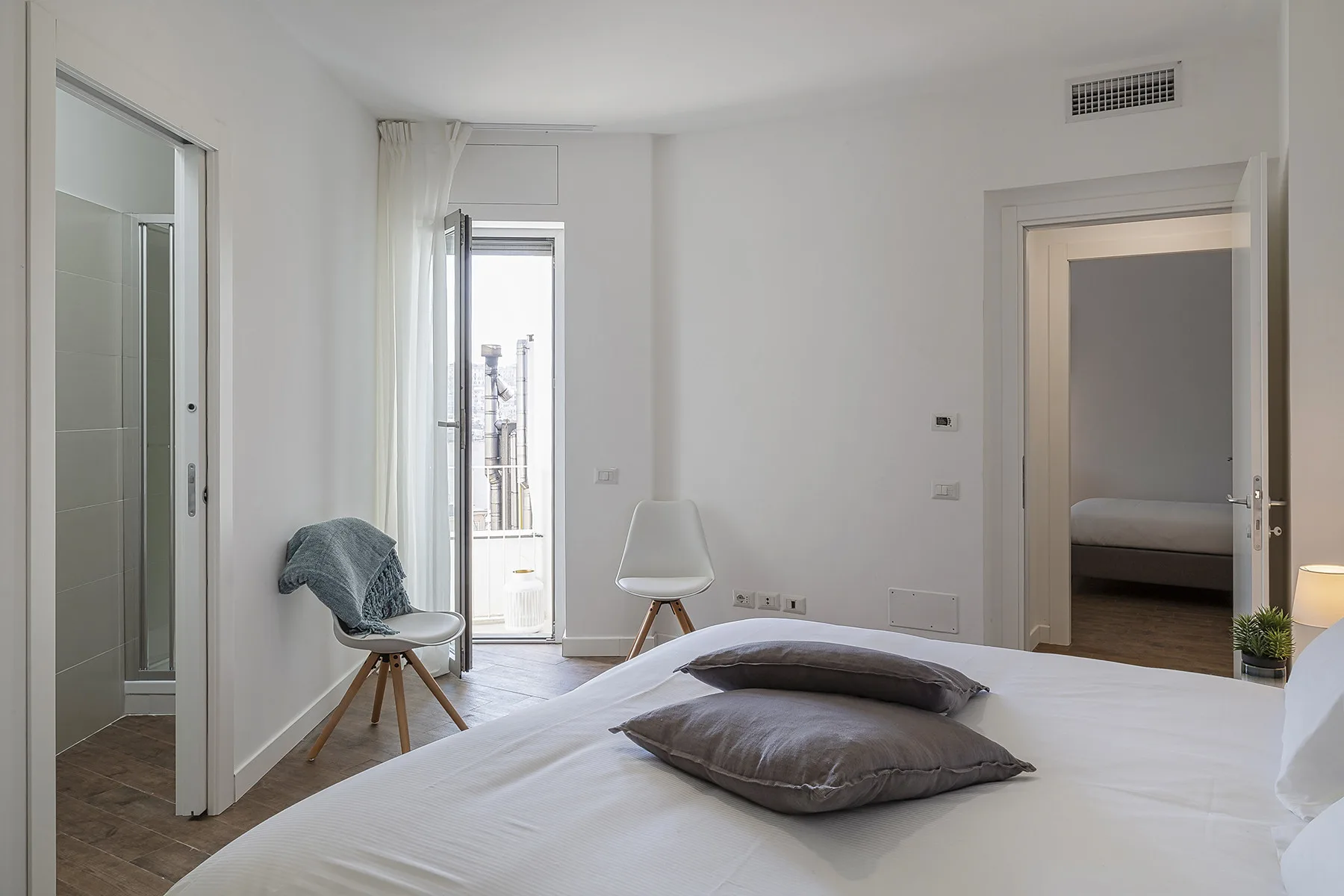 A bright bedroom with a neatly made bed and a chair beside the balcony door.