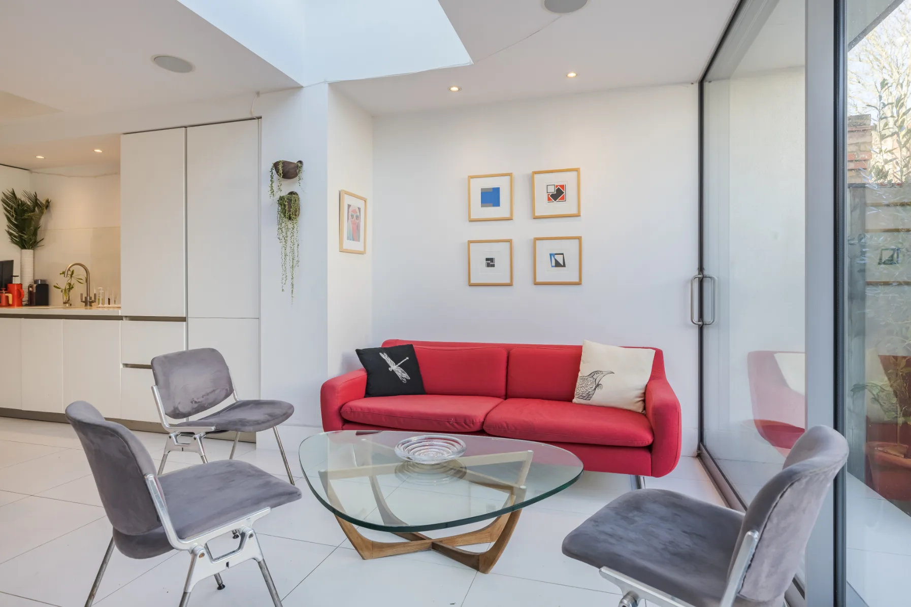 Una sala luminosa con un sofá rojo, una mesa de centro de vidrio y asientos grises.