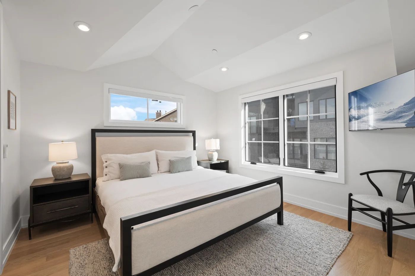 Un dormitorio luminoso con una cama, mesitas de noche y ventanas grandes.