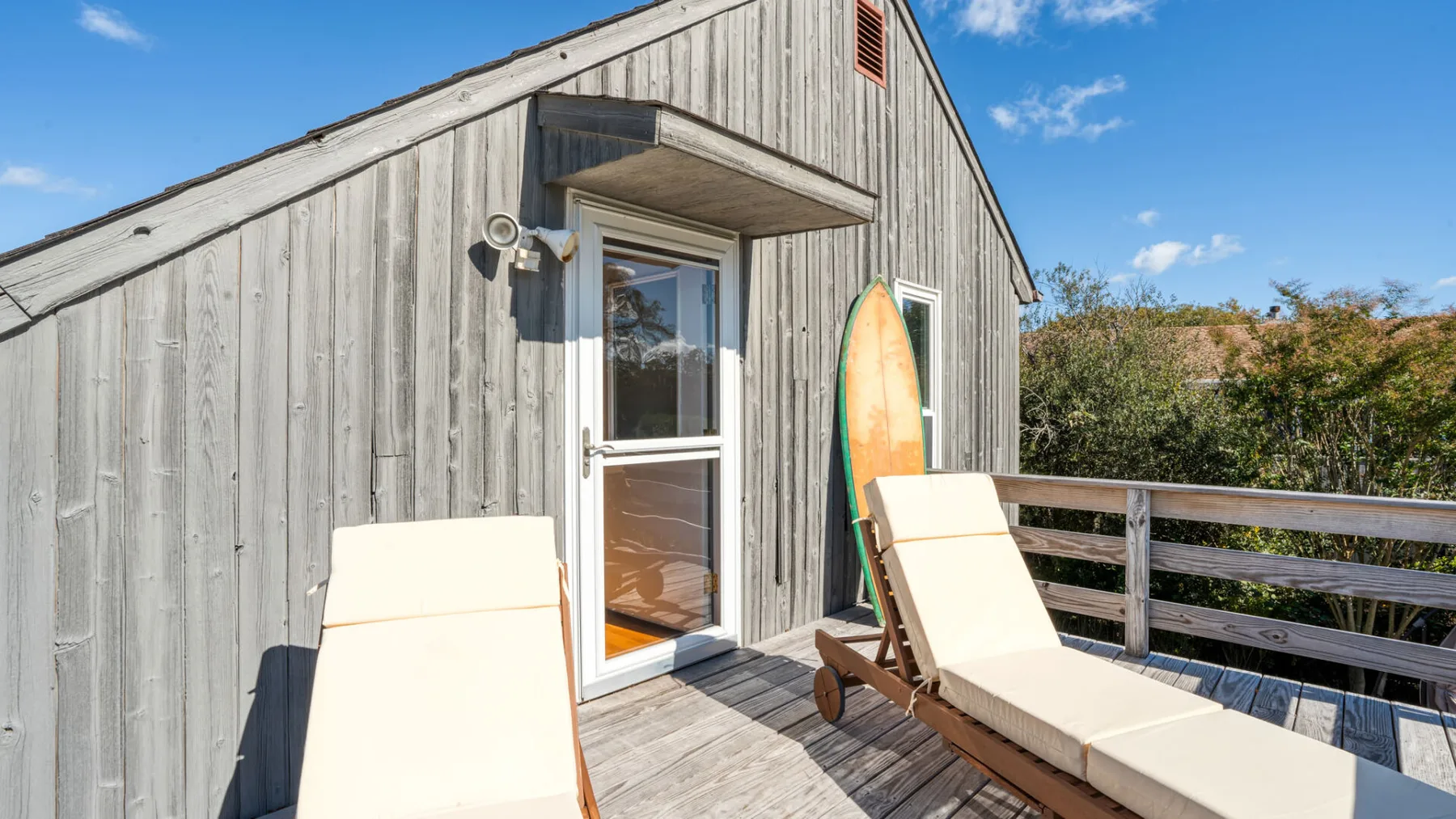 A wooden deck beside a gray cabin has two lounge chairs and a surfboard.