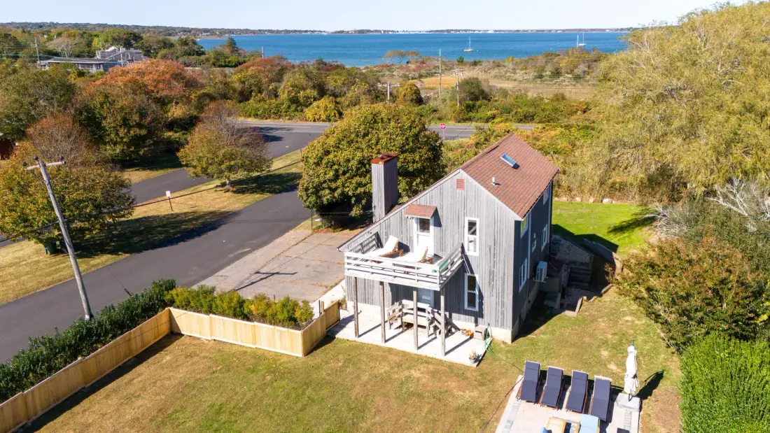 Aerial view of a house with a deck, lawn, and water in the distance.