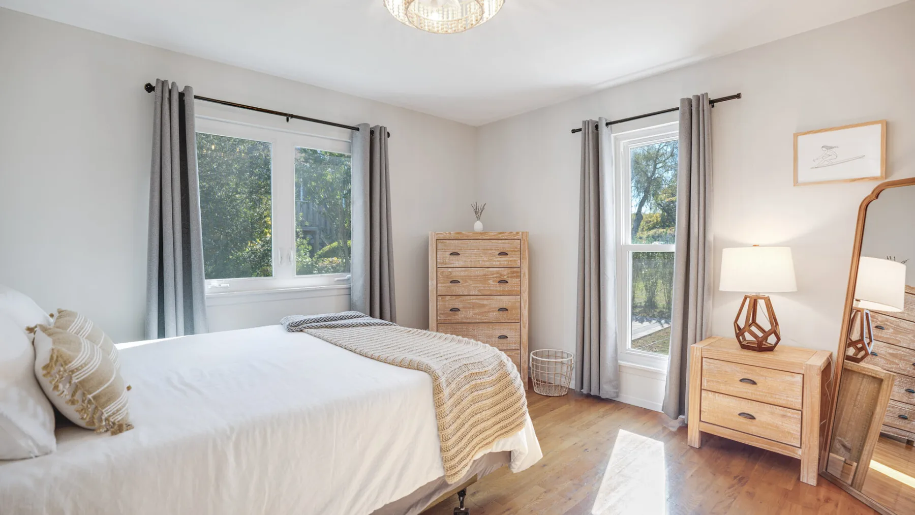 A bright bedroom with a bed, wooden dresser, and windows with gray curtains.