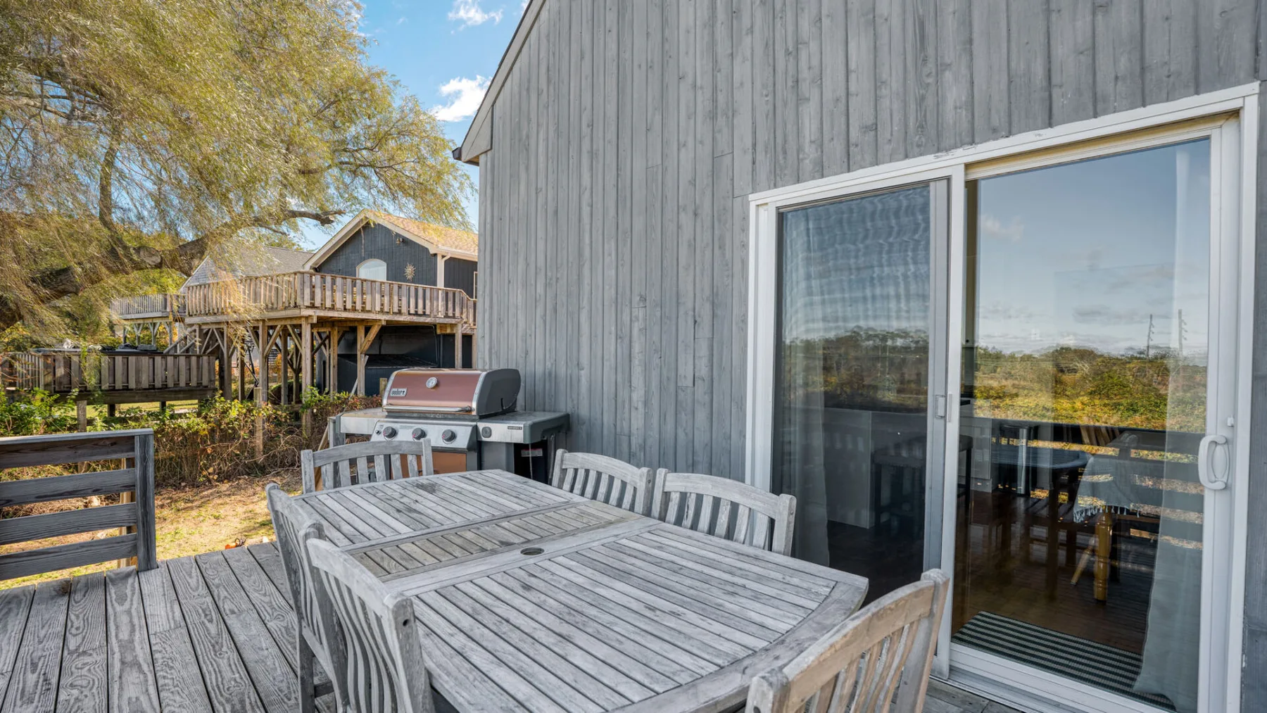 A wooden patio beside a gray-sided building, with outdoor dining furniture and a sliding glass door.