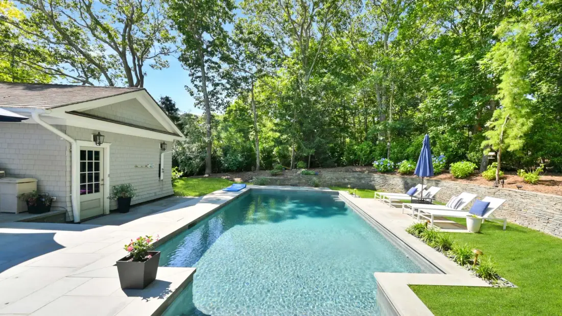 An outdoor swimming pool beside a light-colored building, surrounded by trees and patio seating.