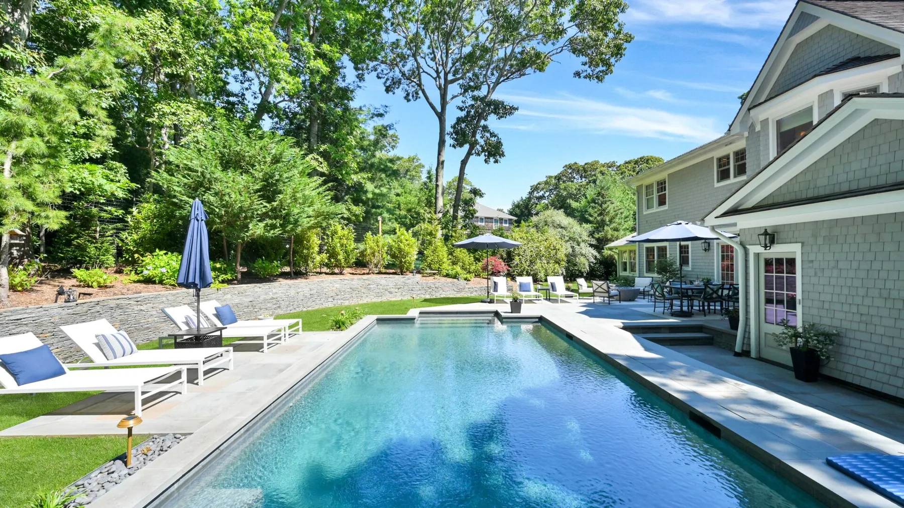 An outdoor swimming pool beside a house, with lounge seating and trees.