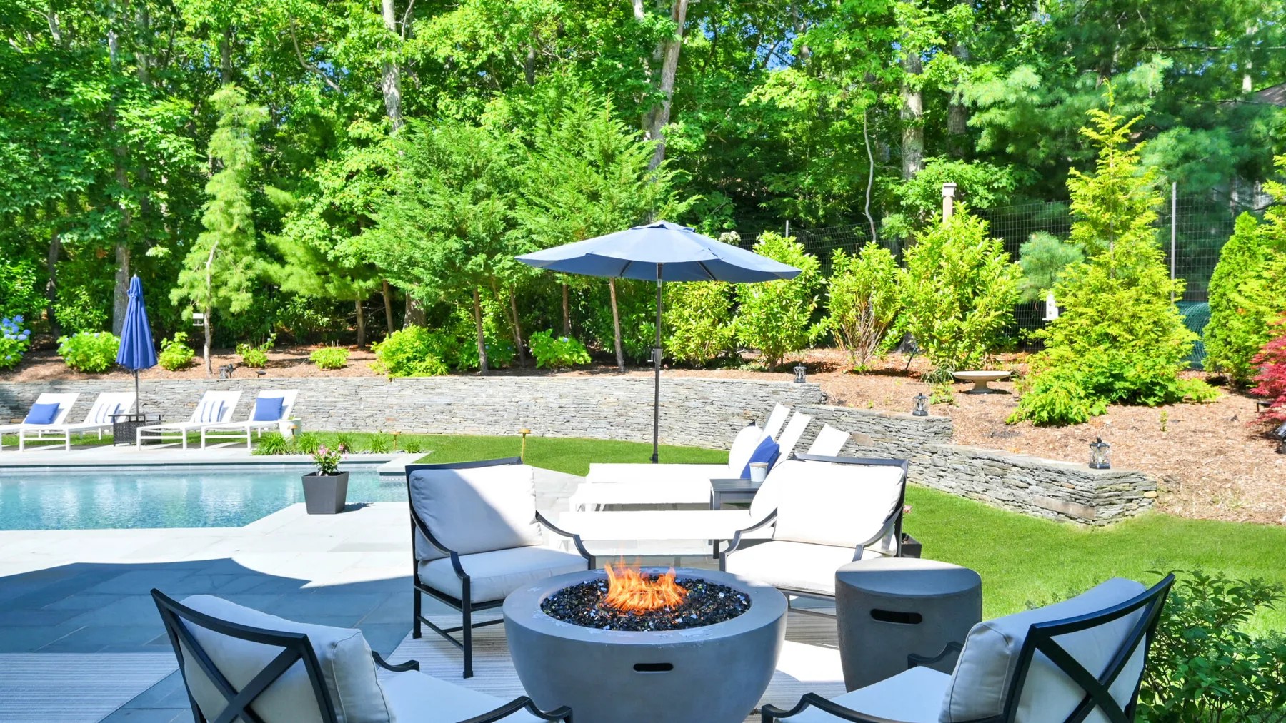 An outdoor patio with a fire pit, seating, and an umbrella beside a pool.