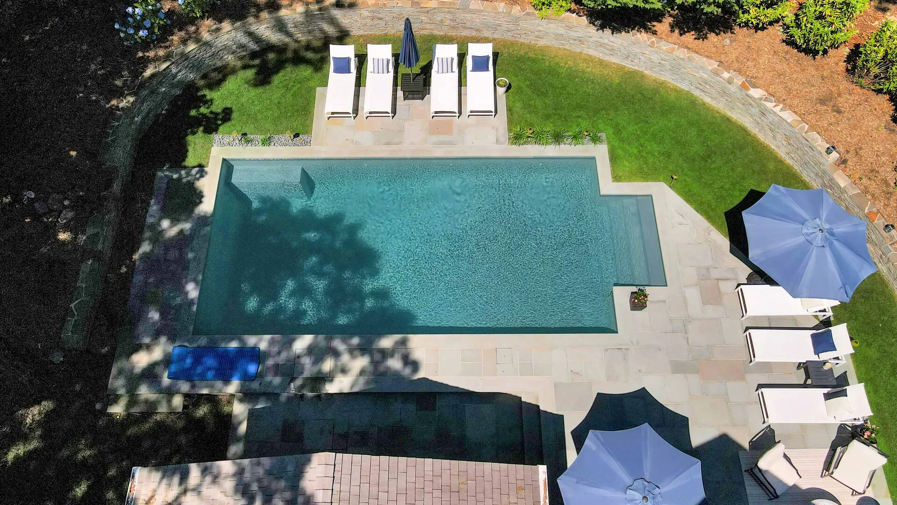 An outdoor swimming pool is surrounded by paved decking, loungers, and umbrellas.