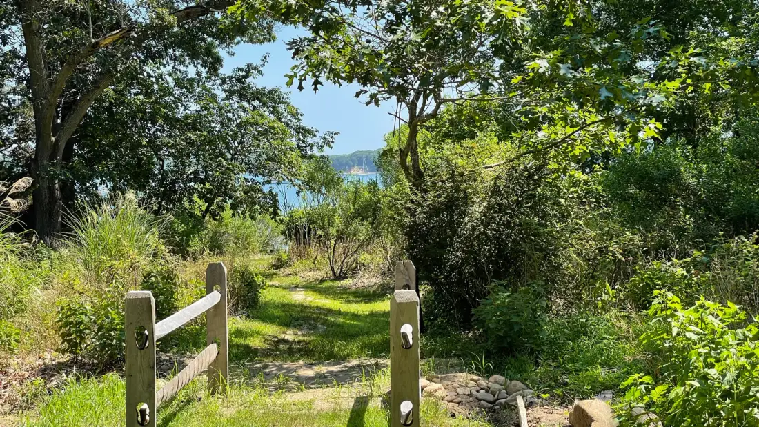 A grassy path passes through a wooden gate toward a glimpse of water.