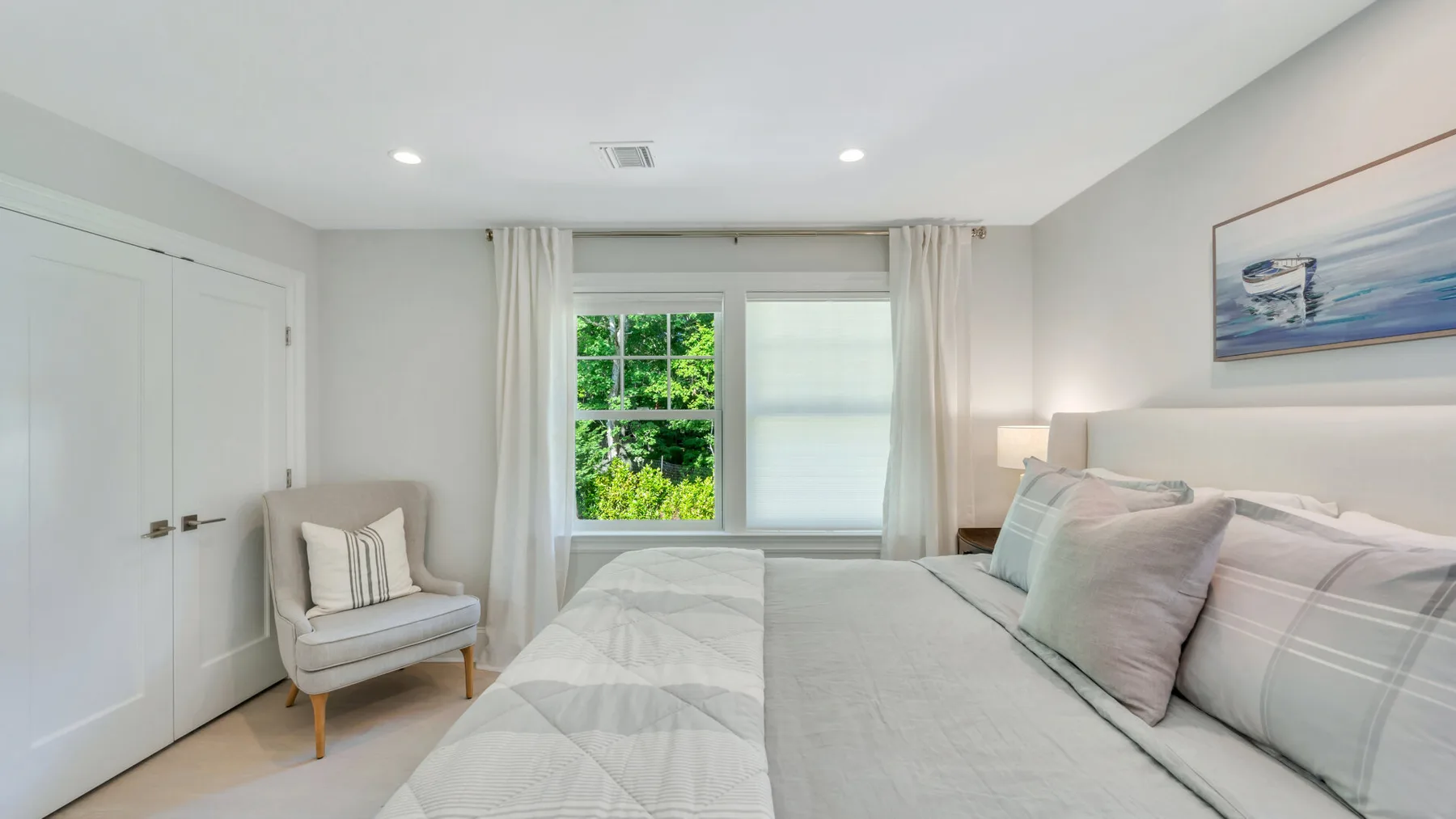 A bright bedroom with a neatly made bed, a window, and an armchair.