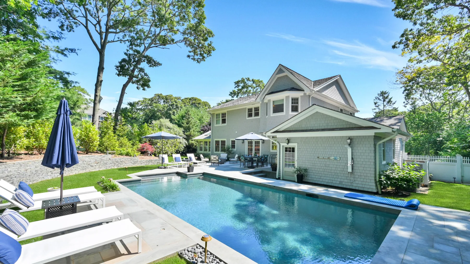 An outdoor swimming pool beside a large house, with lounge seating along the edge.