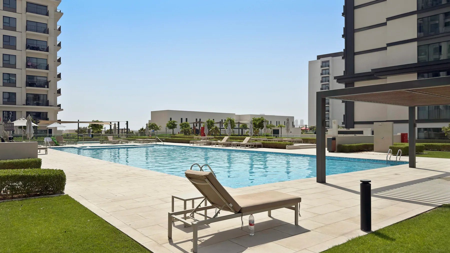 Una piscina al aire libre junto a edificios modernos, con asientos de descanso en la terraza.