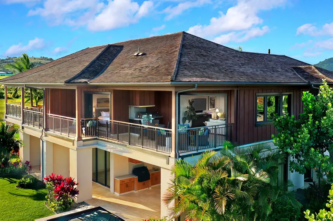 A two-story house with a broad roof, balcony, and surrounding tropical greenery.