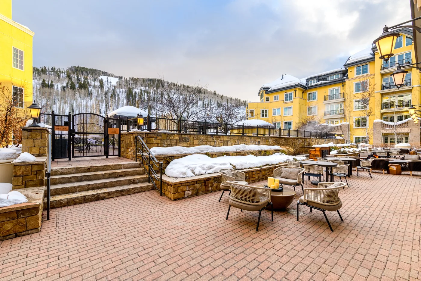 Un patio exterior pavimentado con asientos junto a un edificio amarillo y nieve en las jardineras elevadas.
