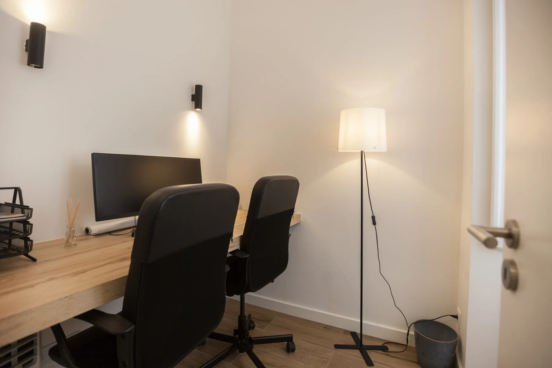 A compact home office has a wooden desk, computer monitor, and two chairs.