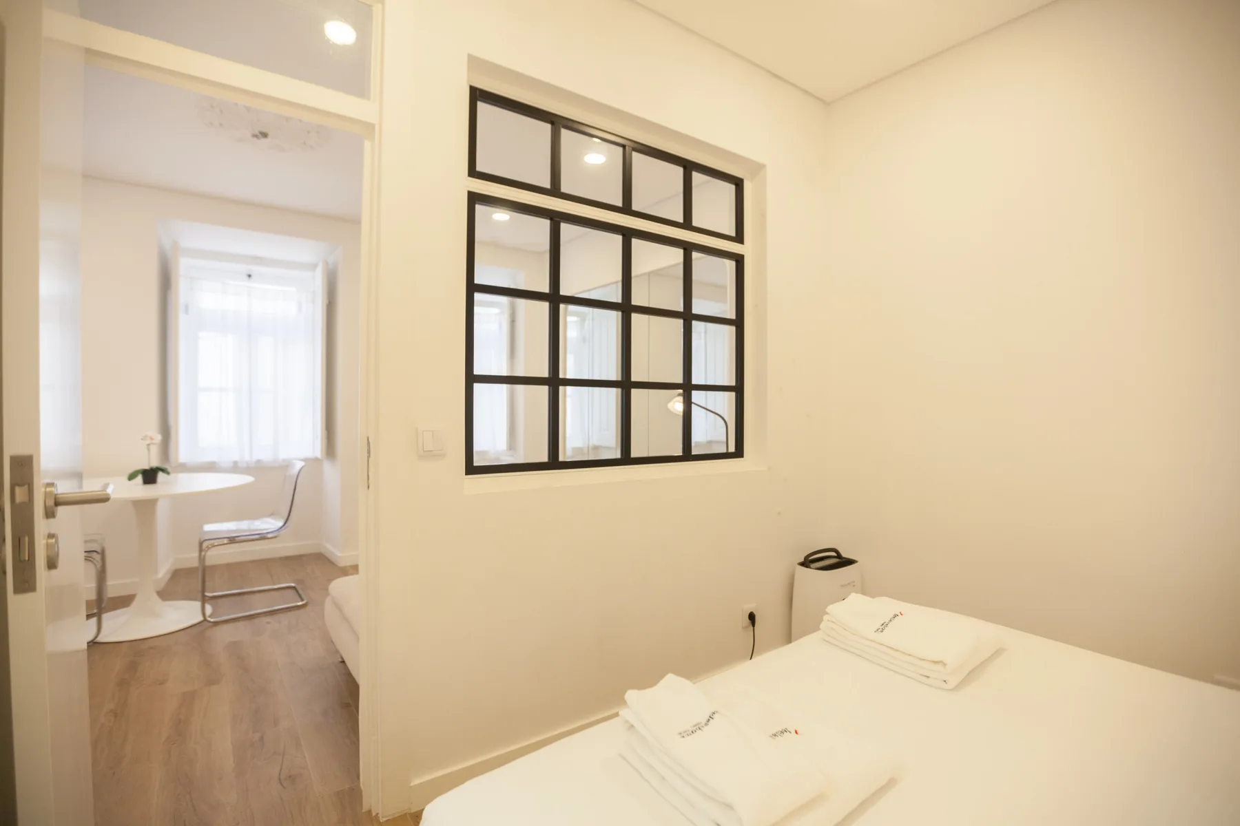 A bright bedroom with neatly made bedding and a black-framed interior window.