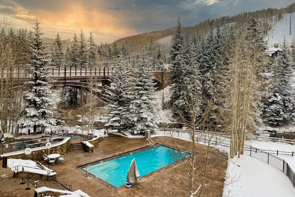 A snowy outdoor pool sits among evergreen trees beside a wooded hillside.