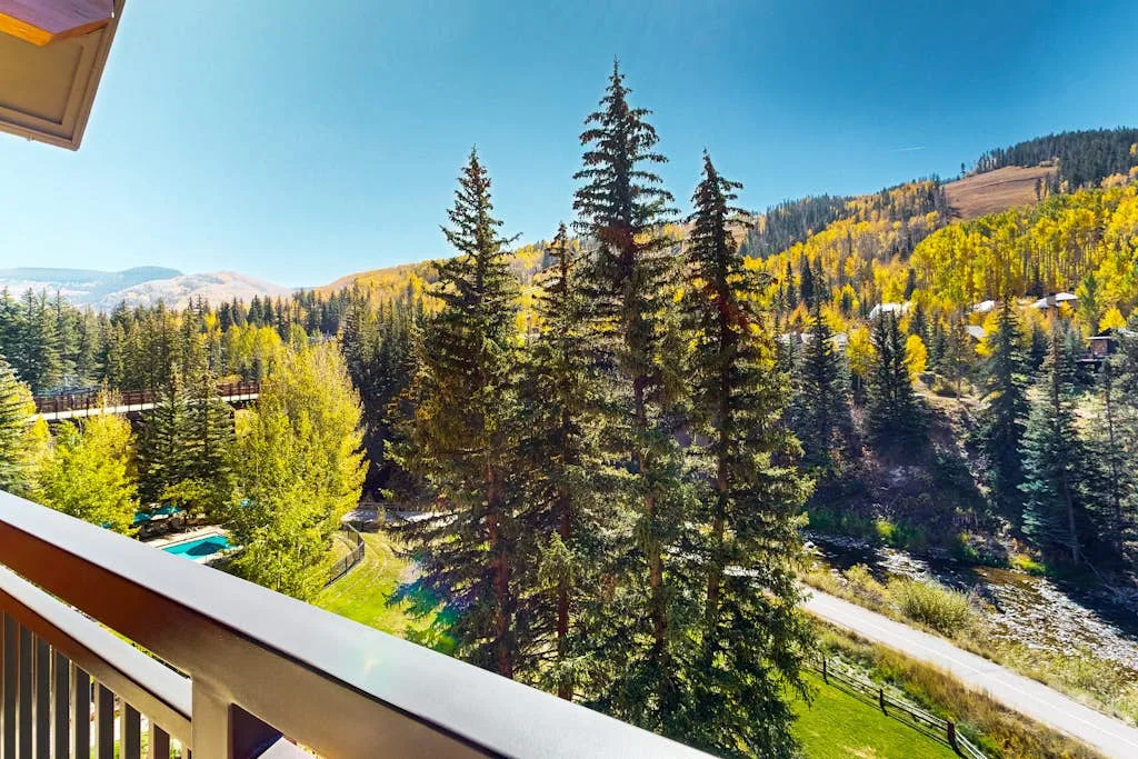 A balcony overlooks evergreen trees, a winding road, and a forested mountain slope.