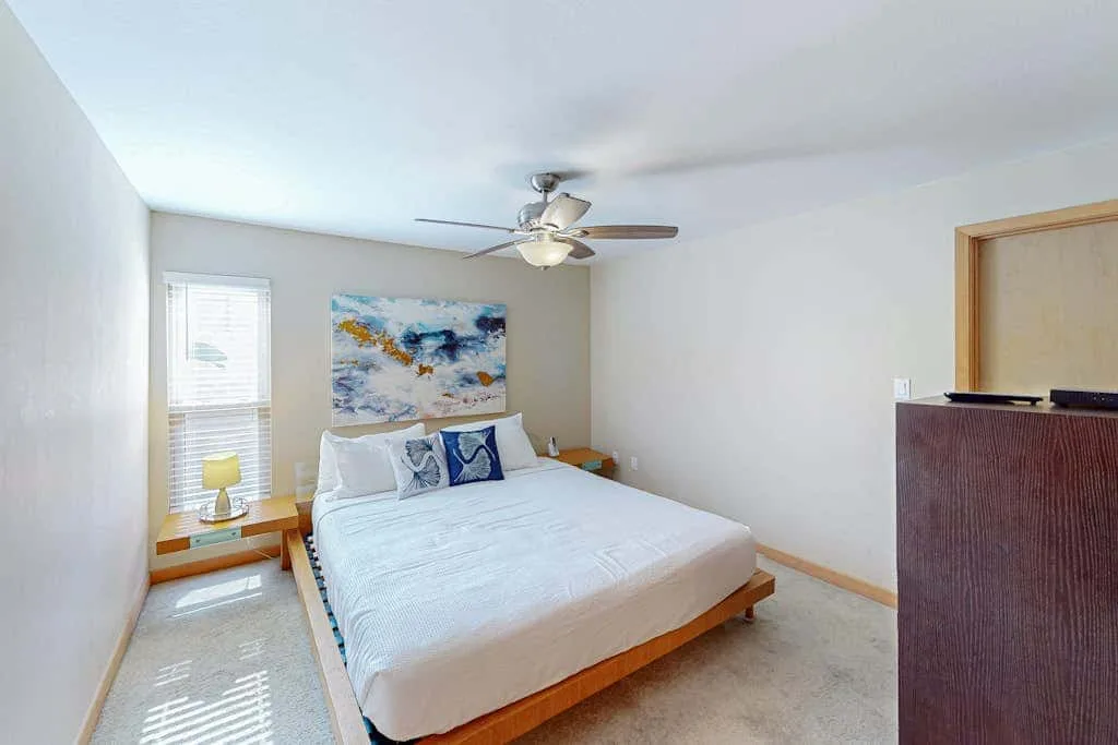 A bedroom with a bed beneath a ceiling fan and a colorful painting on the wall.