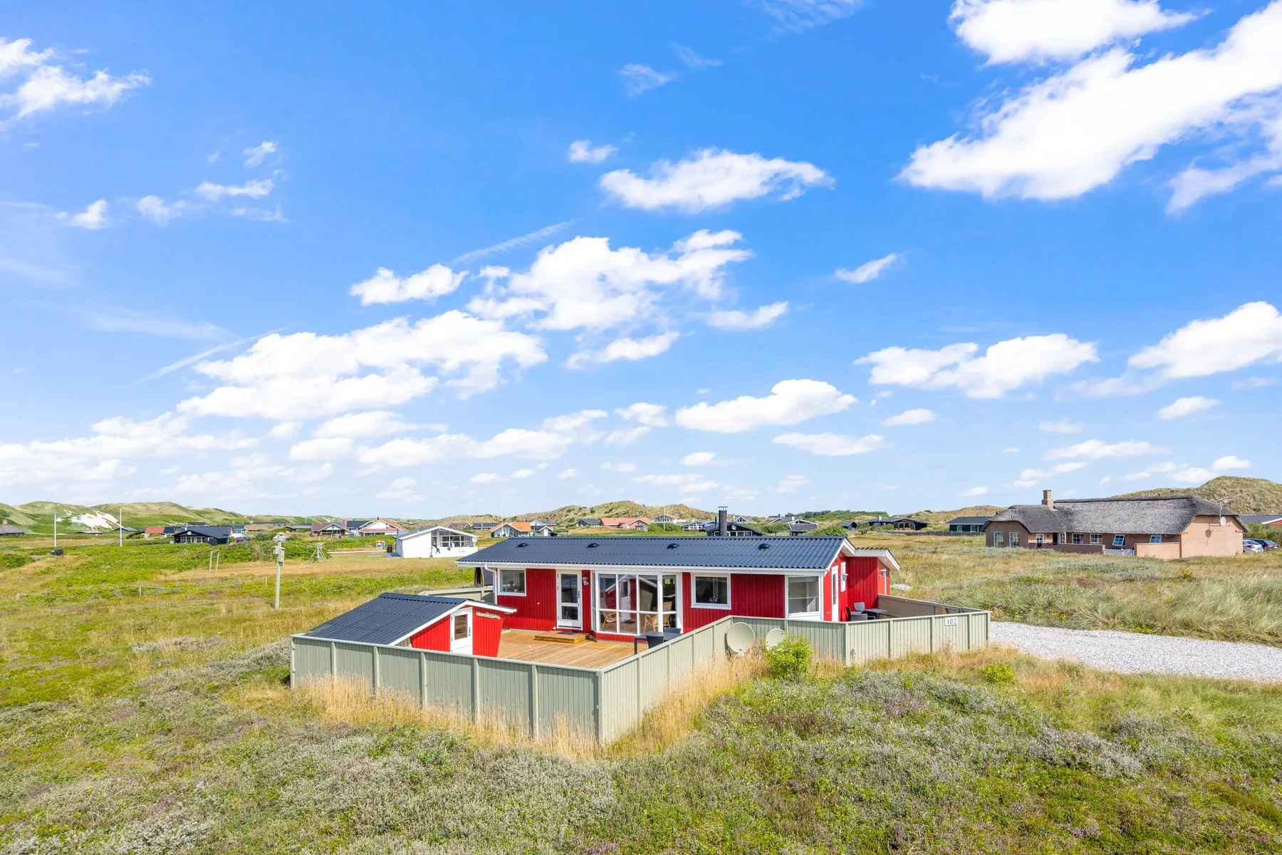 Una casa roja se alza entre terrenos cubiertos de hierba bajo un cielo azul con nubes.