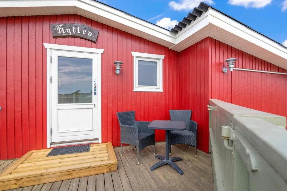 Exterior de una cabaña roja con puerta, ventana y una pequeña zona de asientos en la terraza.