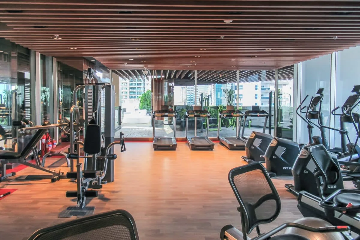 Un gimnasio con máquinas de ejercicio y grandes ventanales con edificios cercanos a la vista.