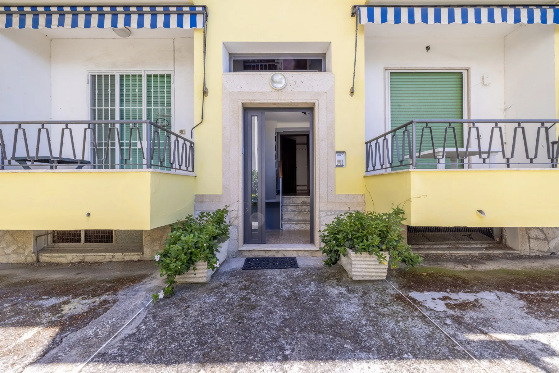 Entrada de un edificio amarillo con dos balcones y arbustos en macetas.