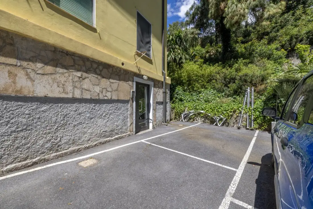 Una zona de estacionamiento al aire libre junto a un edificio, con árboles y vegetación al fondo.