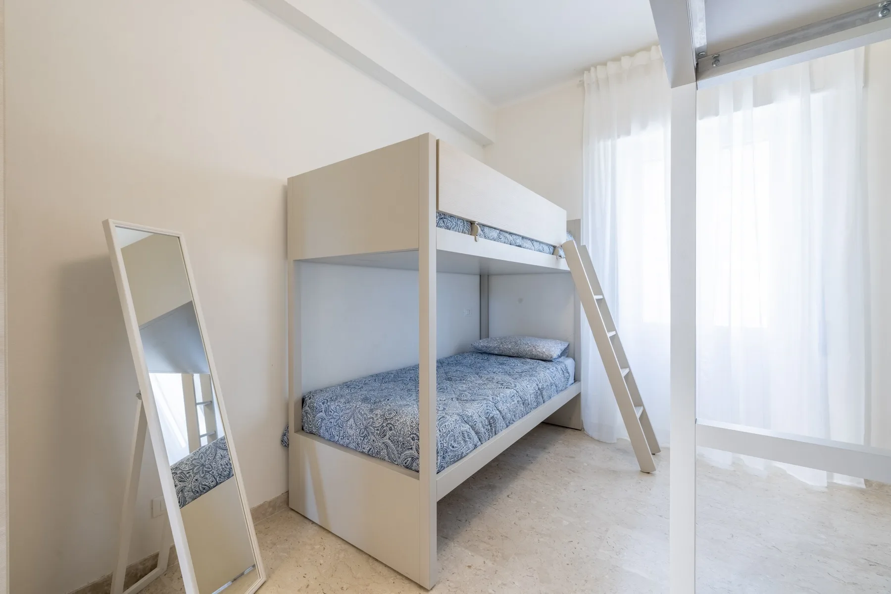 Una litera está junto a una ventana alta con cortinas translúcidas.