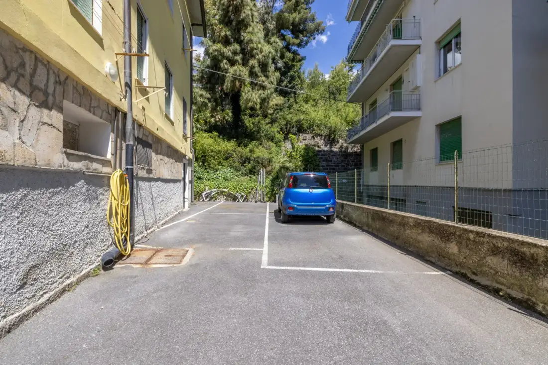 Una zona de estacionamiento pavimentada entre dos edificios, con un coche azul cerca de los árboles.