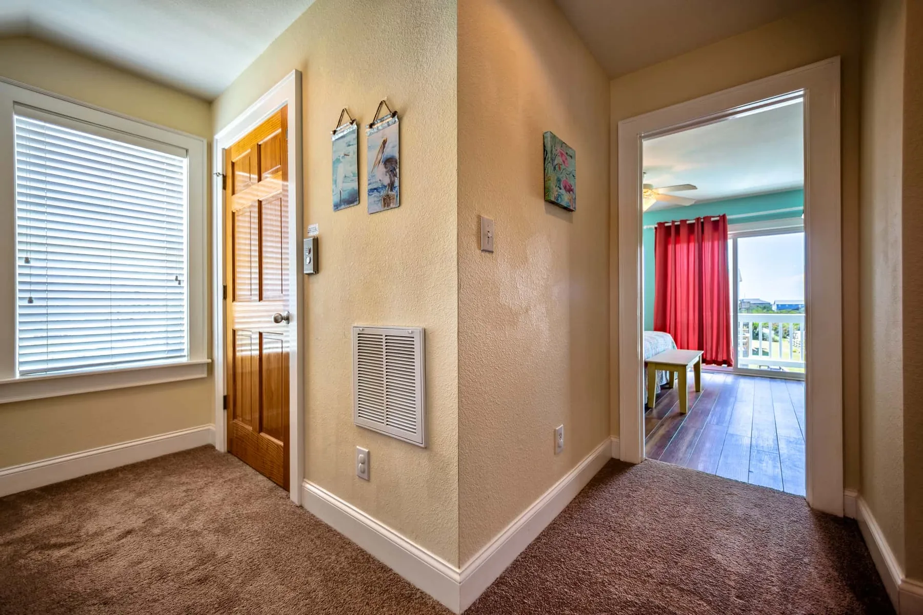 A carpeted hallway with a window, wooden door, and an open doorway to a bright room.