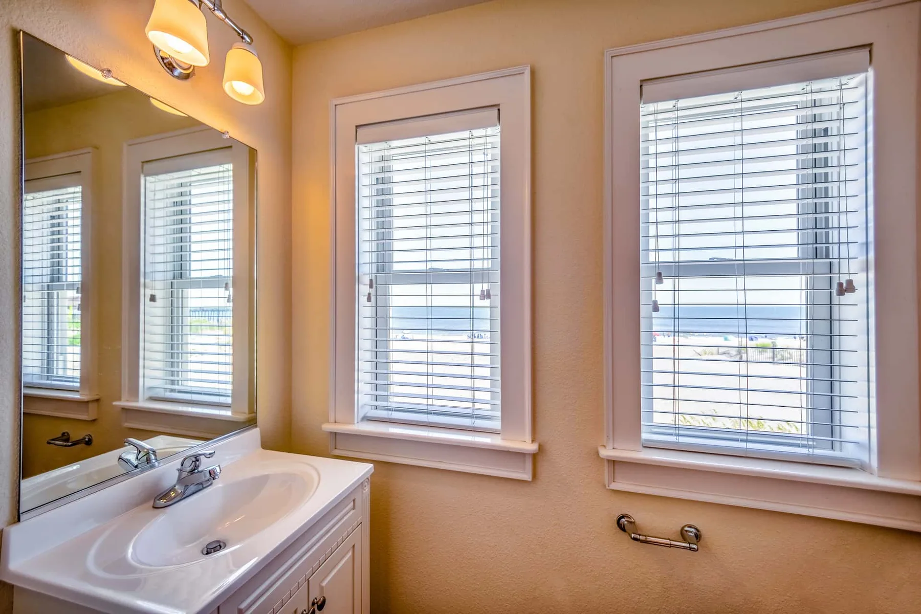 A bathroom vanity sits beneath a wall mirror beside windows with blinds.