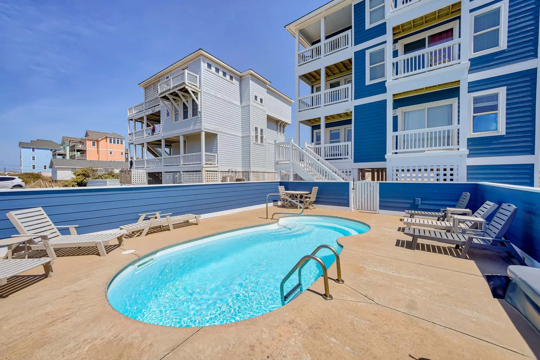 An outdoor swimming pool sits beside a deck with lounge seating and nearby buildings.