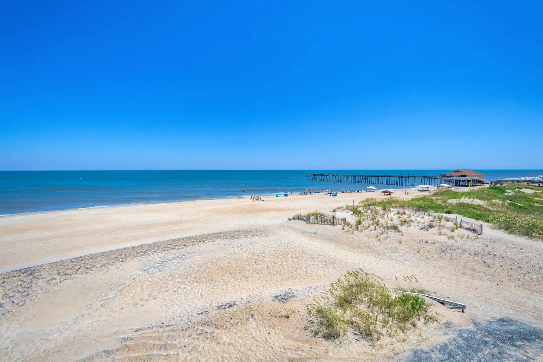 A sandy beach with low dunes, calm water, and a pier in the distance.