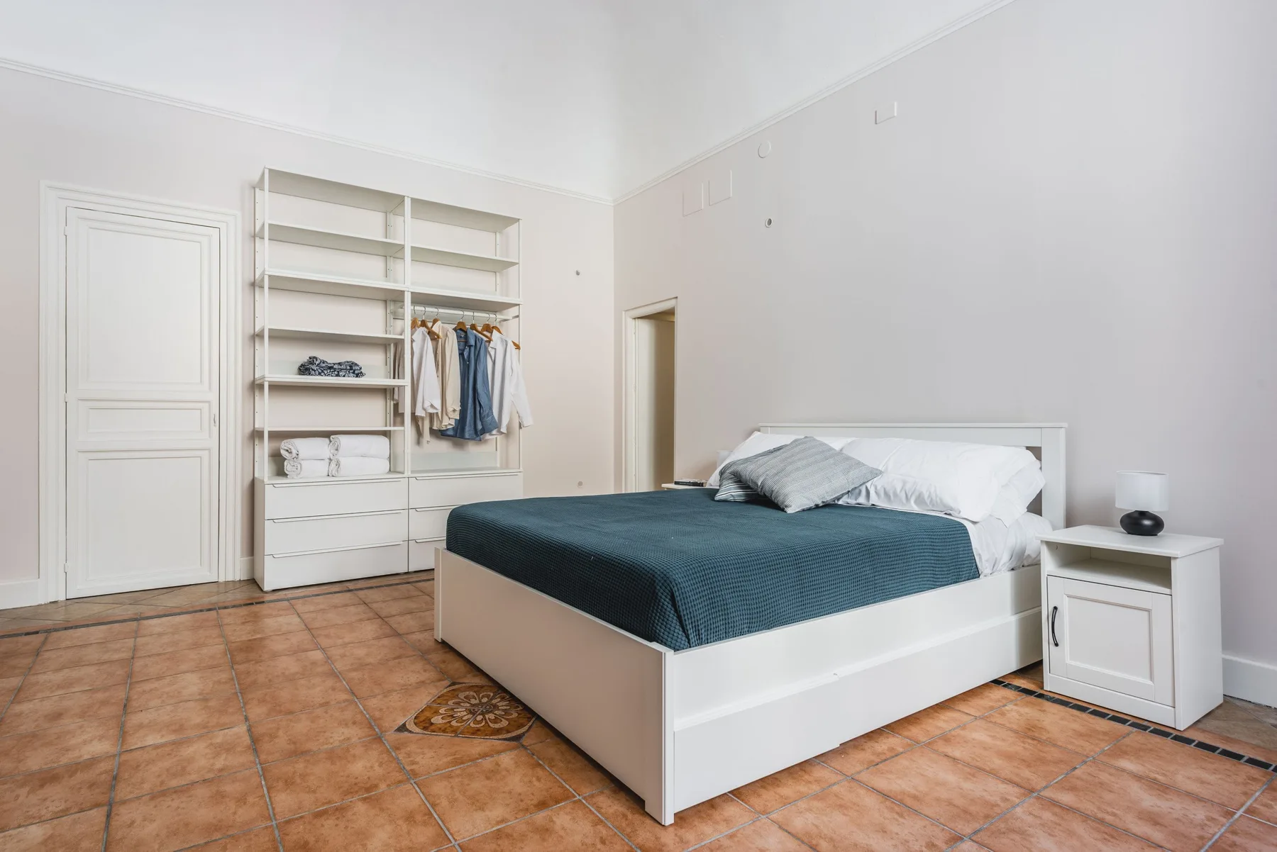 Un dormitorio con una cama blanca, ropa de cama verde azulado oscuro y estanterías abiertas.