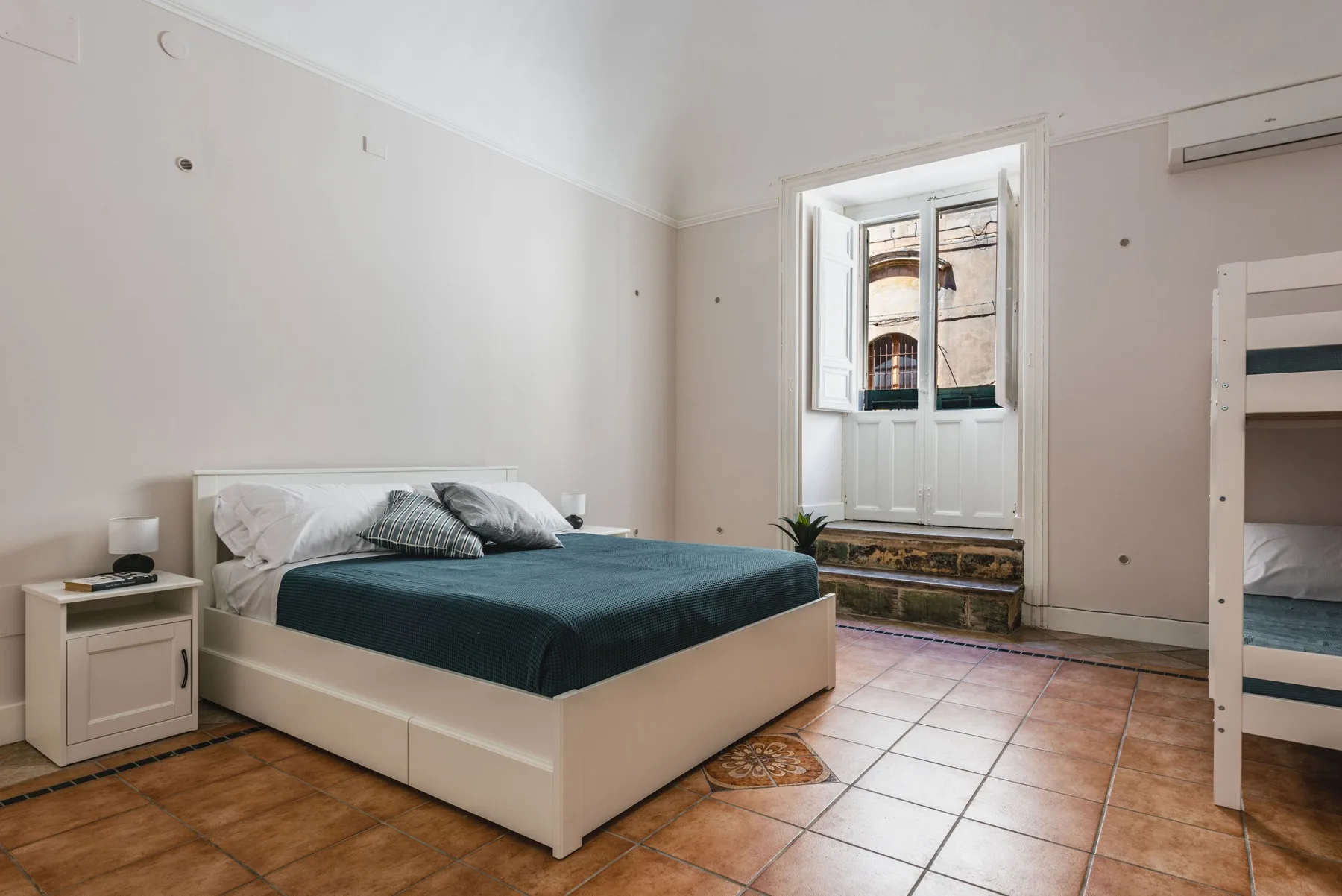 Un dormitorio con una cama, suelo de baldosas y una ventana sobre un alféizar bajo.