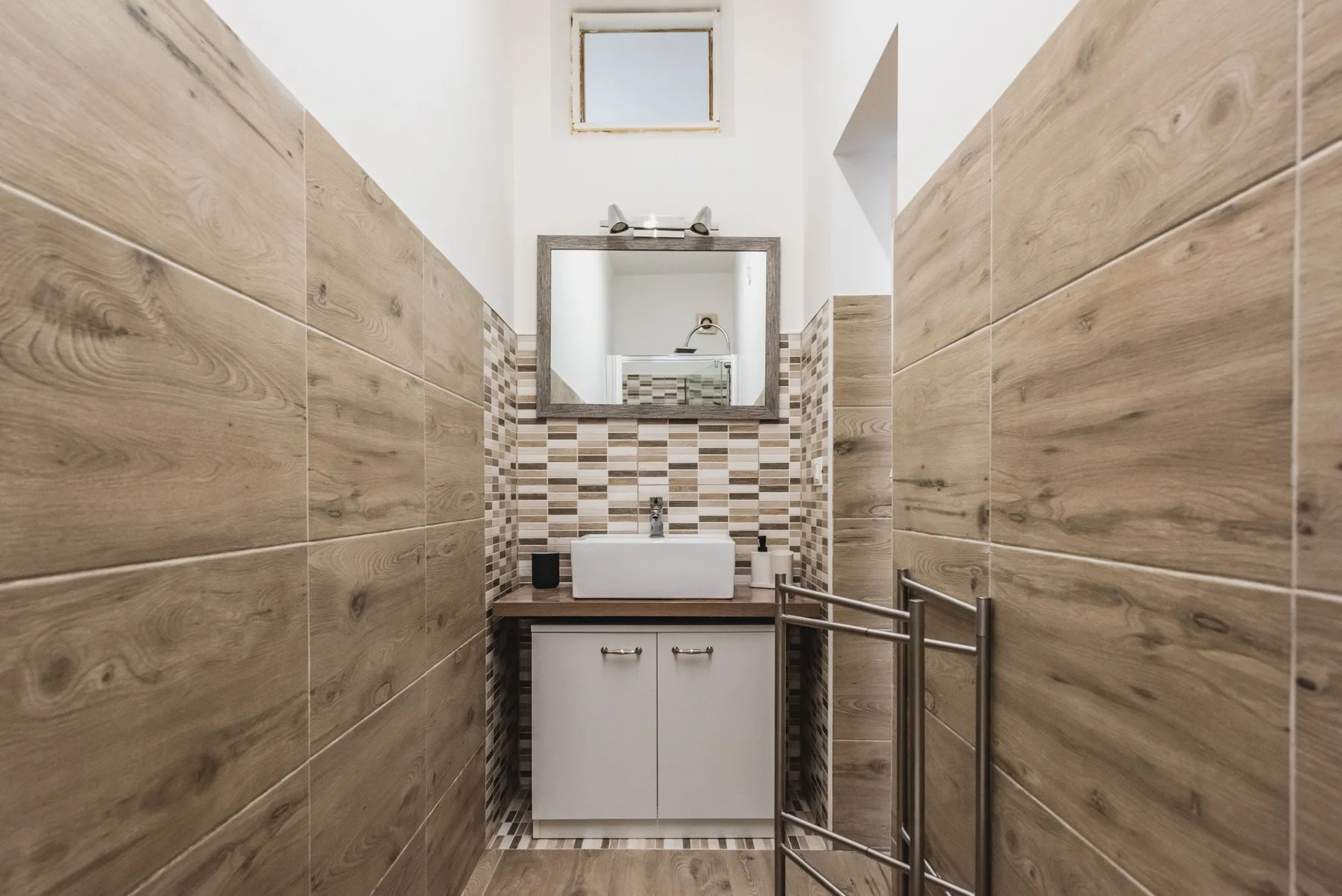 Un baño estrecho con lavabo, espejo y azulejos que imitan la madera.