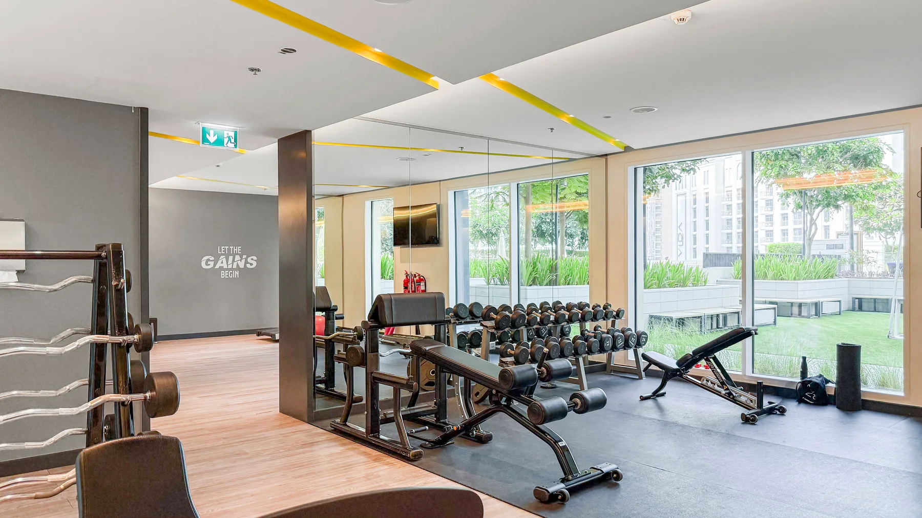 Un gimnasio con estantes de pesas y bancos junto a grandes ventanales.