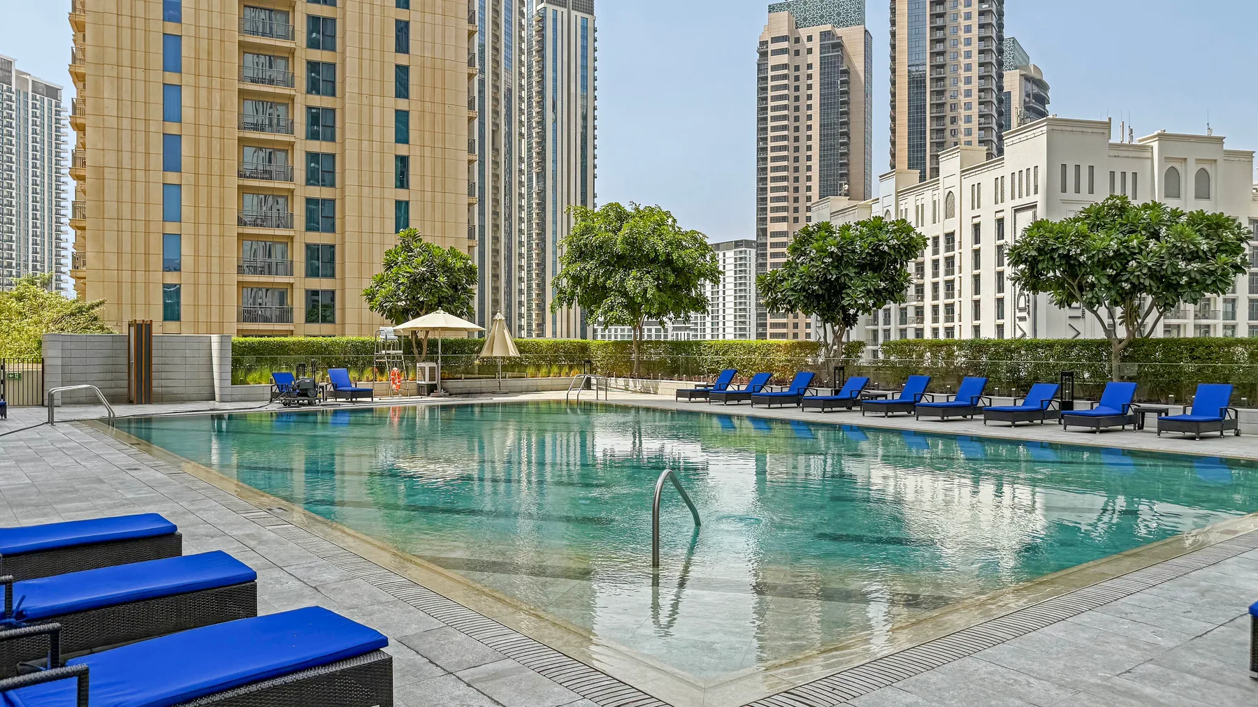 Una piscina al aire libre con tumbonas azules y edificios altos al fondo.
