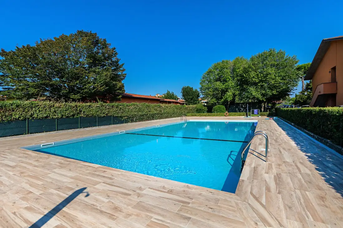 A rectangular outdoor swimming pool is surrounded by a paved deck and greenery.