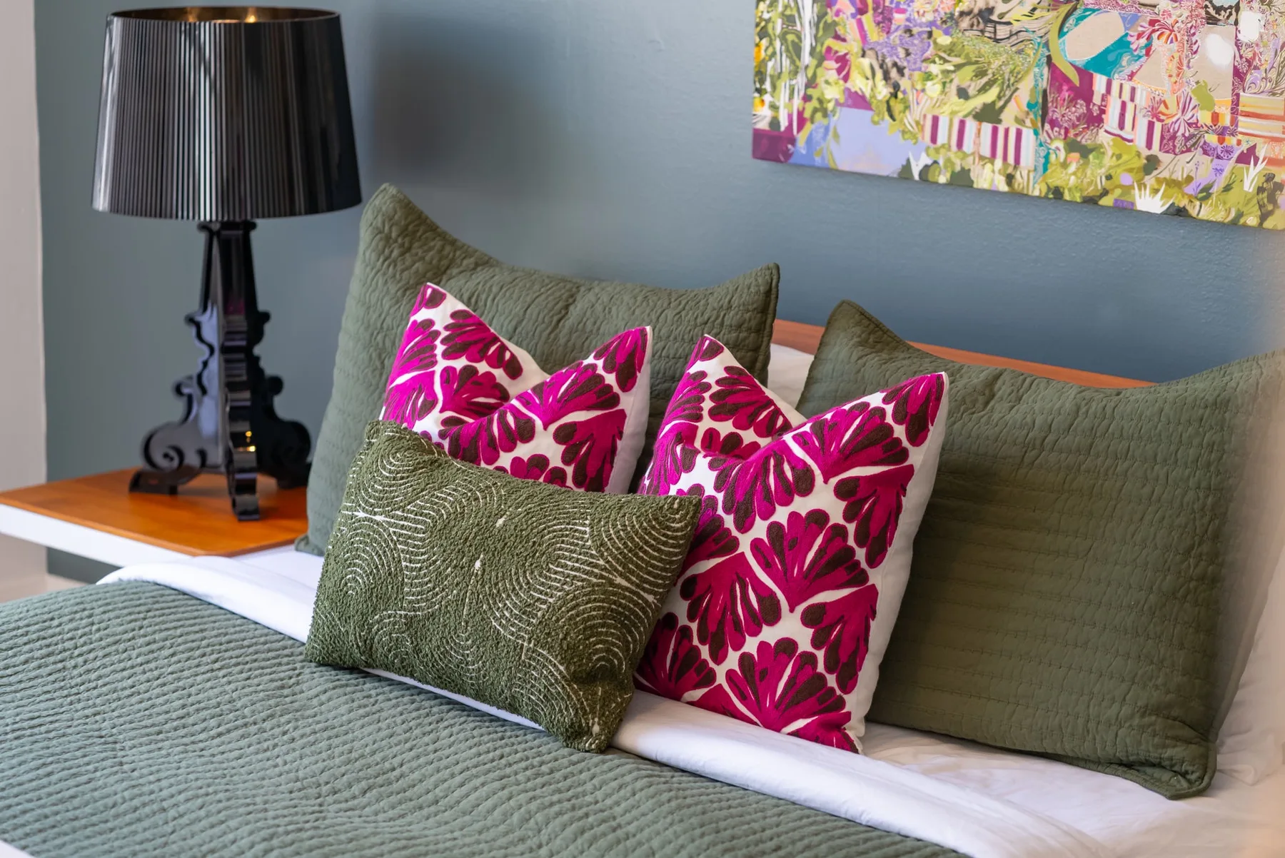 Una cama con ropa de cama verde y cojines estampados de colores está bajo un cuadro.